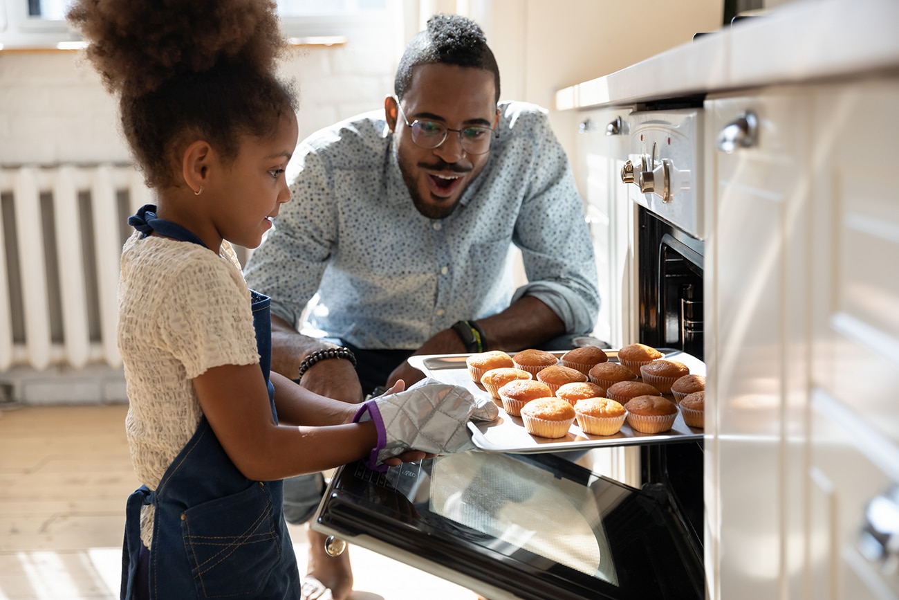 A young girl proudly removes freshly baked muffins from the oven as her smiling father watches in admiration.