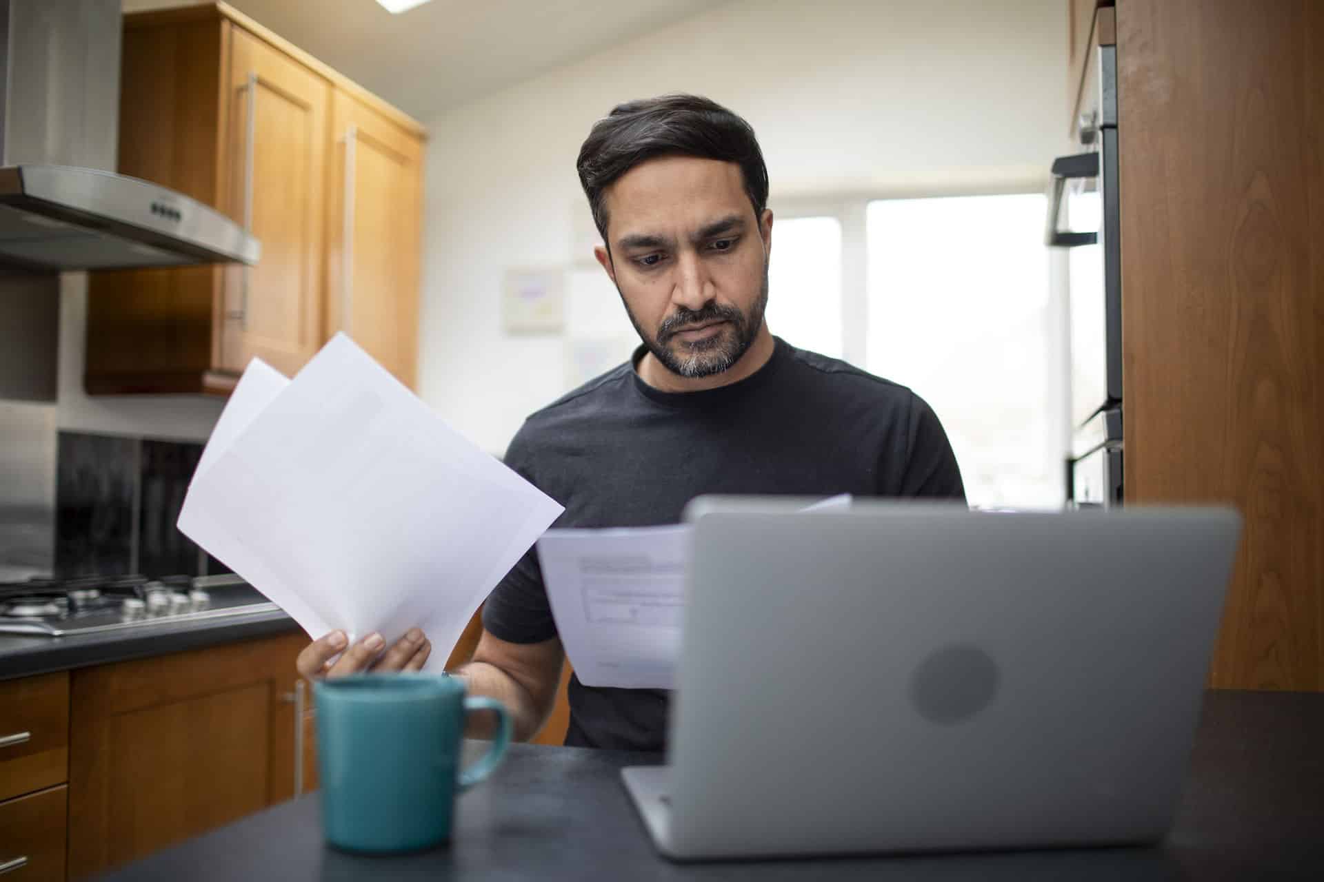 Man sitting at his kitchen table reviewing bills while using a laptop.