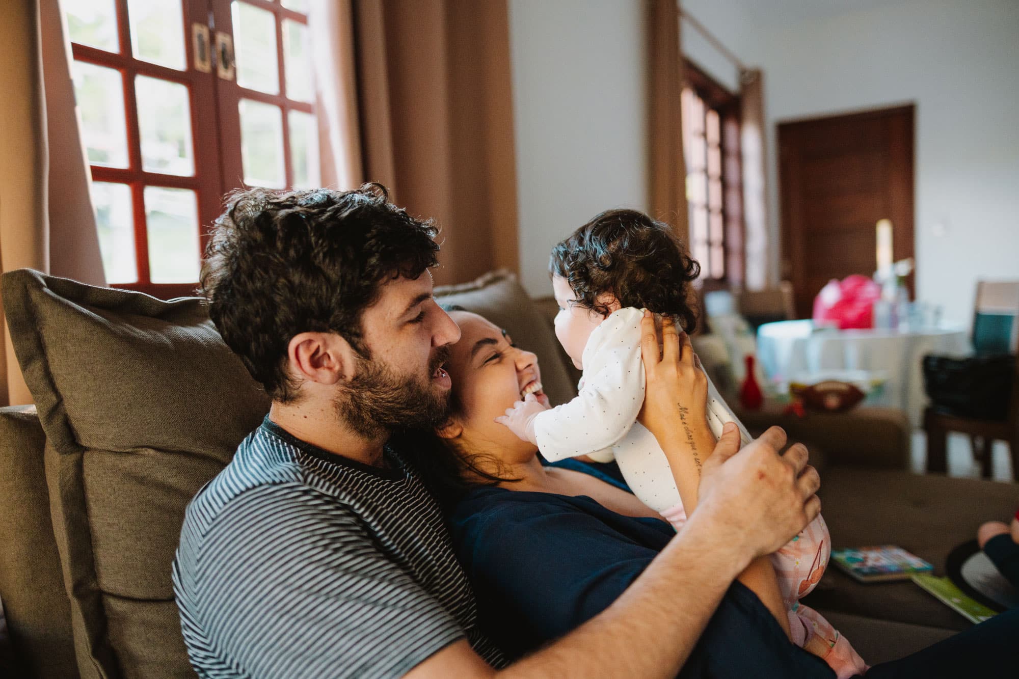 Smiling parents sitting on the couch, holding and playing with their baby in a cozy living room.