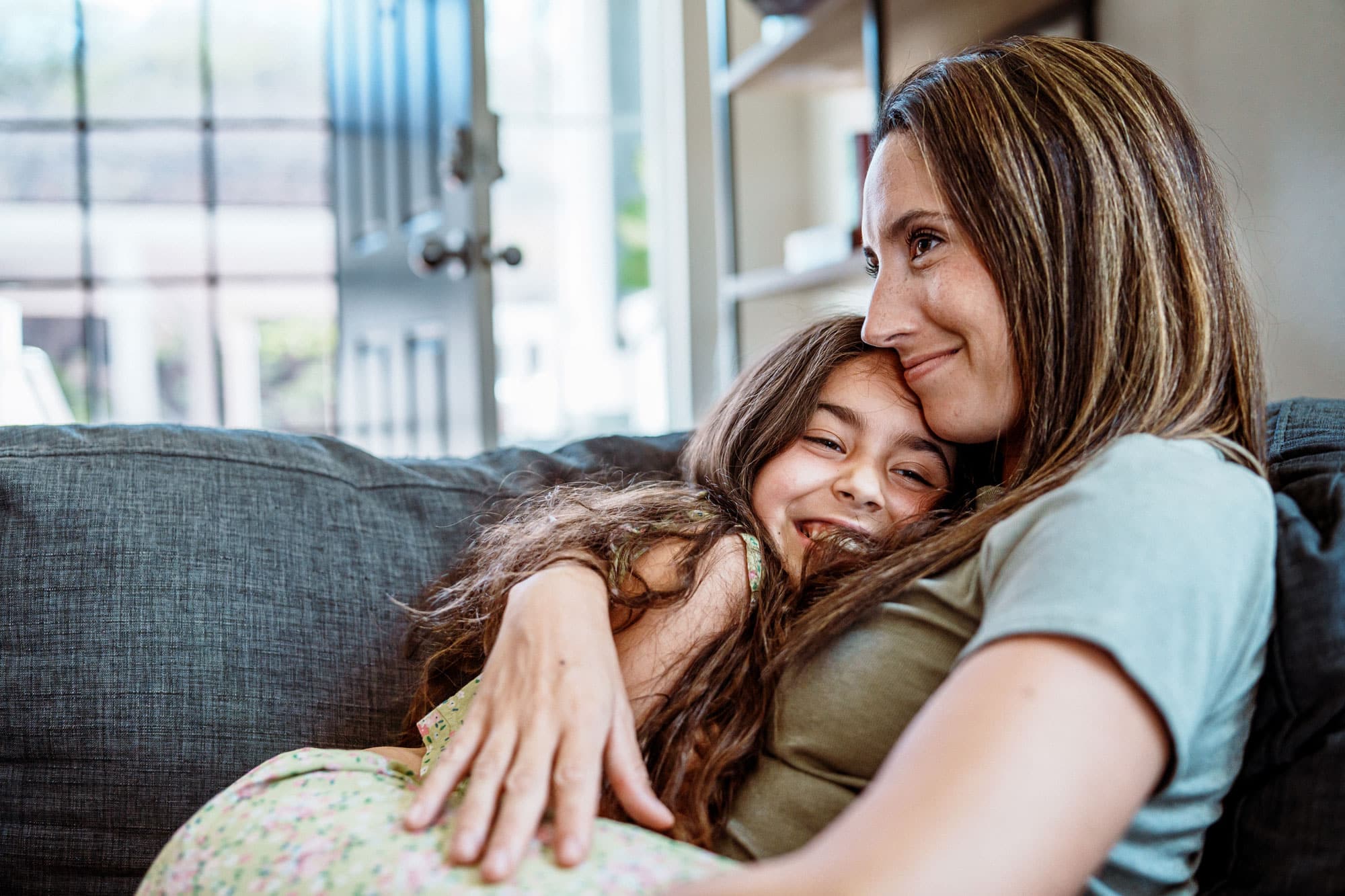 Mother and daughter smiling and hugging on the couch in their home.