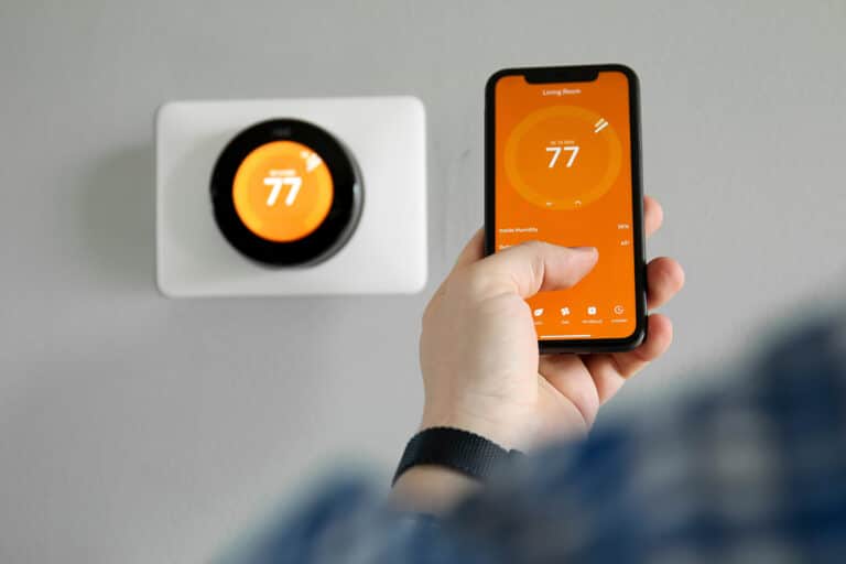 A person adjusts a smart thermostat via a smartphone app. Both thermostat and phone display an orange screen with the number 77, indicating temperature control.