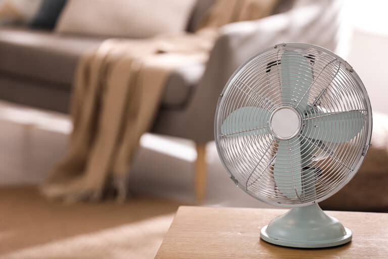 A vintage-style metal fan sits on a wooden table in a cozy living room with a blurred gray couch and beige throw, conveying a warm, comfortable atmosphere.