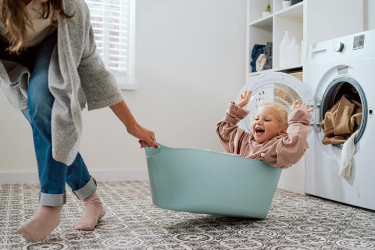 A child joyfully sitting in a laundry basket with arms raised, pulled by a woman in a cozy sweater and jeans. The scene is light-hearted and playful.